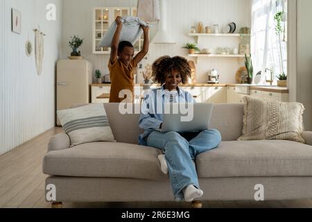 Un petit fils attaque une femme souriante, joue avec un oreiller. Mère travaille sur un ordinateur portable, petit enfant distrait Banque D'Images