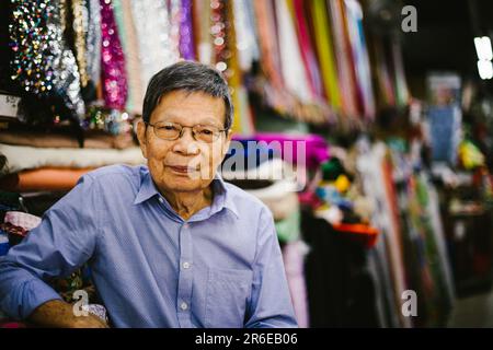 Un homme asiatique plus âgé vend des tissus dans son magasin en lunettes Banque D'Images