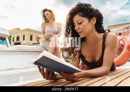 Une femme caucasienne aux cheveux bouclés est engrosée en lisant sur le pont du bateau, tandis que son ami brésilien est assis en arrière-plan, en lisant également. Ils enjo Banque D'Images