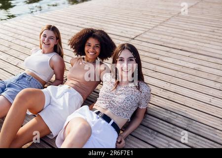 Les adolescentes souriantes et multiraciales se trouvent sur un sol en bois à l'extérieur. Rire jolies filles de différentes nationalités dans les vêtements décontractés ont grand Banque D'Images