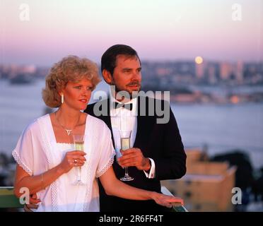 Couple dans des vêtements de couture buvant du champagne sur la terrasse sur le toit donnant sur un port. Banque D'Images