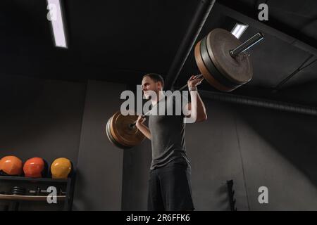 Homme de forme physique faisant de l'entraînement de poids avec barbell. Jeune athlète musclé ayant l'entraînement à la salle de gym sombre. Poids de levage de carrossier faisant des coudossier. Concept CrossFit Banque D'Images