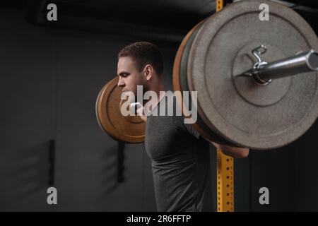 Portrait d'un bodybuilder de sexe masculin faisant des squats dans le dos en salle de sport. Musclé beau jeune homme se préparer à faire des squats barbelés portant une chemise noire. CrossFit et concept d'entraînement Banque D'Images