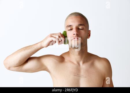 Soulèvement du visage, soin anti-rides. Jeune homme beau faisant un massage du visage avec un outil cosmétologique sur fond blanc de studio Banque D'Images