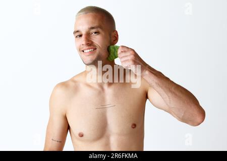 Jeune homme beau faisant un massage du visage avec un outil cosmétologique sur fond blanc de studio. Levage de face. Circulation Banque D'Images
