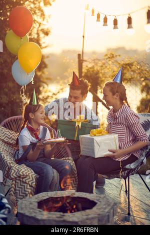 Fête d'anniversaire. Maman et papa donnant des cadeaux à la fille d'anniversaire, assis autour d'une cheminée à l'extérieur dans un chalet confortable. Célébration, style de vie conc Banque D'Images