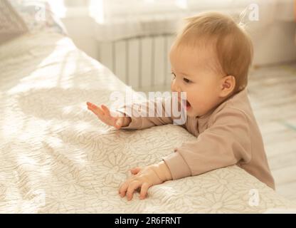 Une fille asiatique se tient dans la chambre à coucher, s'appuyant sur le lit et essayant de prendre ses premiers pas. Banque D'Images