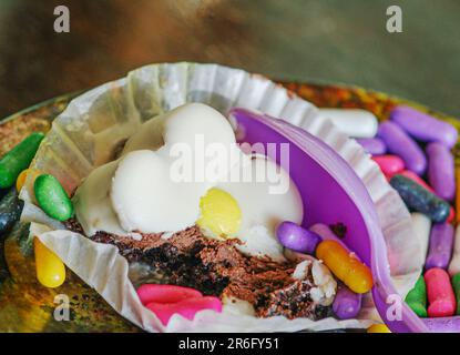 Un dessert partiellement mangé est dans un porte-gâteau avec des bonbons colorés. Le dessert est un gâteau au chocolat au caramel de souris avec une cuillère en plastique mauve. Banque D'Images