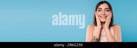 positivité, tatouée jeune femme avec des cheveux courts dans le débardeur haut sourire et toucher son visage sur fond bleu, tenue décontractée, la mode de gen z, le bonheur Banque D'Images