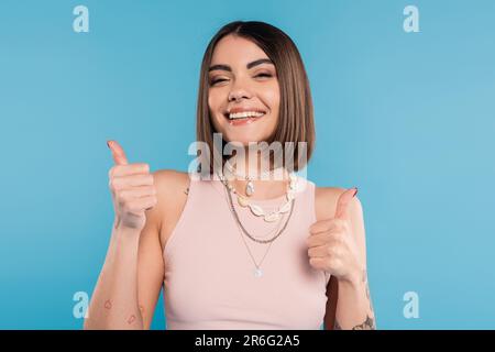 positivité, tatouée jeune femme avec de courts cheveux brunette dans le débardeur sourire et montrant les pouces sur fond bleu, tenue décontractée, mode gen z, Banque D'Images