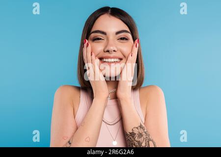 positivité, tatouée jeune femme avec de courts cheveux de brunette dans le débardeur haut sourire et toucher son visage sur fond bleu, tenue décontractée, mode gen z, Banque D'Images