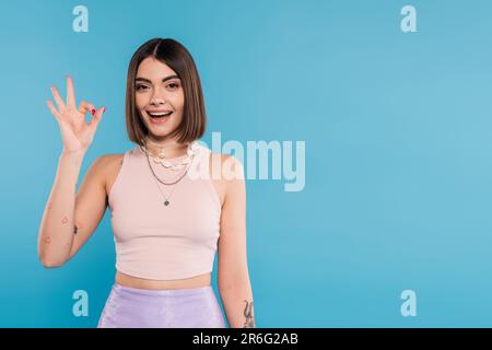 excitante, tatouée jeune femme avec des cheveux courts dans le débardeur haut sourire et montrant le geste ok sur fond bleu, tenue décontractée, la mode de gen z, fashiona Banque D'Images