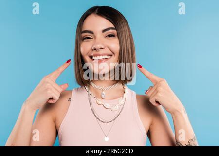 excitation, tatouée jeune femme avec de courts cheveux de brunette dans le débardeur haut sourire et pointant vers ses joues sur fond bleu, tenue décontractée, gen z fash Banque D'Images