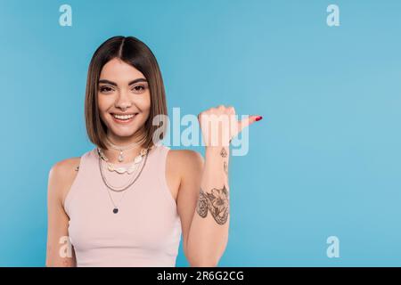 gaieté, tatouée jeune femme avec de courts cheveux de brunette dans le débardeur haut souriant et pointant avec le pouce sur fond bleu, tenue décontractée, gen z fashi Banque D'Images
