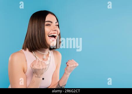 excitation, tatouée jeune femme avec de courts cheveux de brunette dans le débardeur haut sourire et gesturant avec les mains sur fond bleu, tenue décontractée, gen z fashio Banque D'Images