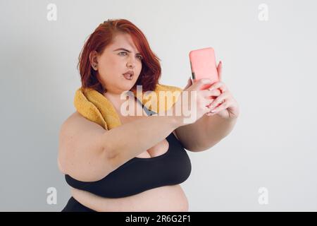 jeune femme latine d'origine argentine portant des vêtements de sport noirs et de grande taille prenant une photo de selfie avec son téléphone, debout sur fond blanc Banque D'Images