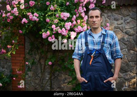 Beau jardinier masculin dans la tenue de jardinage, regardant la caméra, debout à son lieu de travail dans l'arrière-cour d'un manoir Banque D'Images