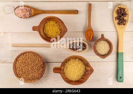 Céréales sèches et assaisonnements dans des tasses en bois avec des cuillères en bois sur une table en bois, macro, vue de dessus. Banque D'Images