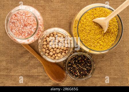 Céréales sèches et assaisonnements dans un bol en verre avec cuillères en bois sur toile de jute, macro, vue du dessus. Banque D'Images