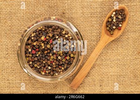 Grains de poivre noir dans un récipient en verre avec une cuillère en bois sur un chiffon de jute, macro, vue de dessus. Banque D'Images