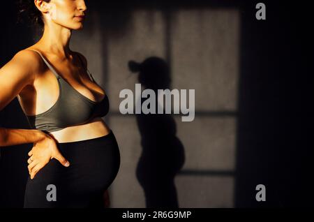 Femme enceinte contre un mur de texture grise avec silhouette d'ombre. Une femme attend un bébé debout sur les côtés et touchant le ventre avec ses mains. Mothe Banque D'Images