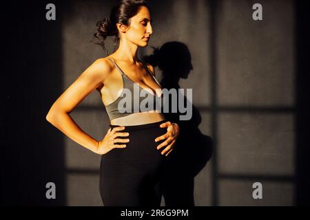 Femme enceinte contre un mur de texture grise avec silhouette d'ombre. Une femme attend un bébé debout sur les côtés et touchant le ventre avec ses mains. Mothe Banque D'Images