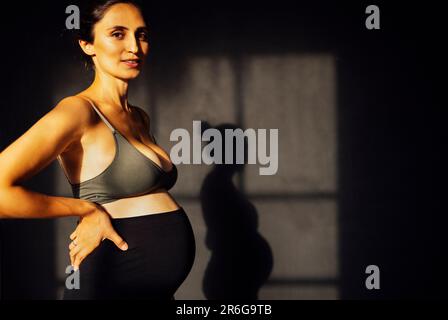 Femme enceinte contre un mur de texture grise avec silhouette d'ombre. Une femme attend un bébé debout sur les côtés et touchant le ventre avec ses mains. Mothe Banque D'Images