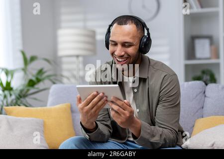 Jeune homme hispanique souriant assis sur un canapé à la maison, portant un casque et utilisant une tablette. Banque D'Images