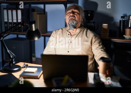 Homme d'âge moyen avec des cheveux gris travaillant au bureau la nuit regardant endormi et fatigué, épuisé pour la fatigue et la gueule de bois, les yeux paresseux le matin. Banque D'Images