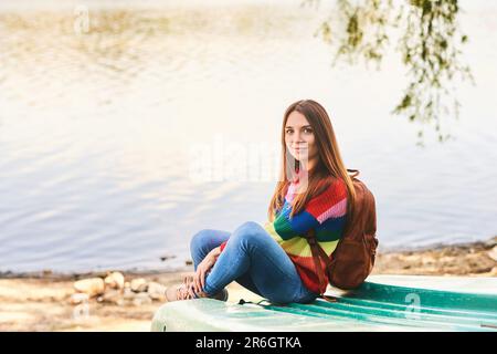 Portrait extérieur de la jeune belle femme se reposant au bord de la rivière lors d'une belle journée chaude, portant un pull-over coloré et un sac à dos Banque D'Images