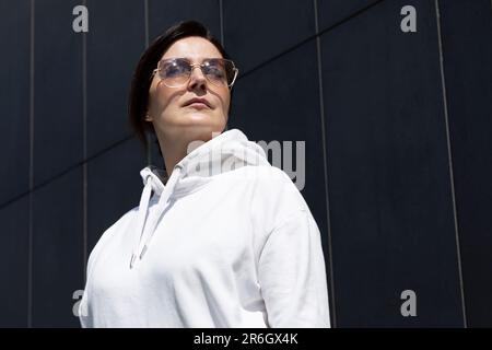 Portrait jolie femme d'âge mûr Brunette en blanc sweat à capuche et lunettes de soleil regarde loin, Dark Background Wall. Sportive confiant 40 Yo magnifique caucasien Banque D'Images