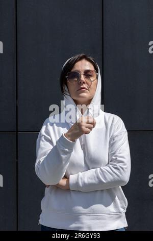 Portrait une femme brune mûre avec capuche sur la tête, porte un sweat à capuche blanc et des lunettes de soleil a l'air droit, mur de fond sombre. Sportive confiant, 40 ans Banque D'Images