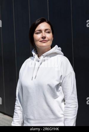 Portrait souriant femme d'âge mûr Brunette en blanc sweat à capuche regarder loin, Dark Background Wall. Sportive détendue 40 Yo belle femme caucasienne dans la ville Banque D'Images