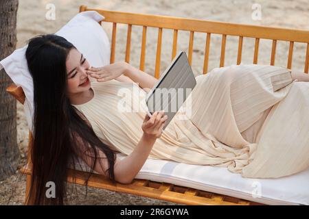 Jeune femme se reposant sur un banc sur une plage de sable et regardant un nouvel épisode de sitcom sur un ordinateur tablette Banque D'Images