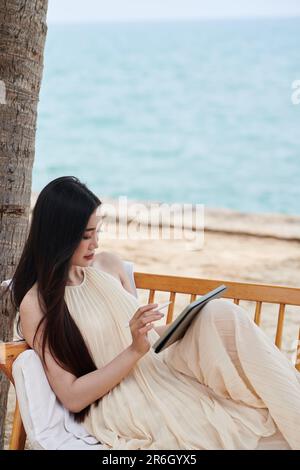 Jeune femme en robe de soie passant du temps sur la plage et le contrat de relecture sur ordinateur de tablette Banque D'Images