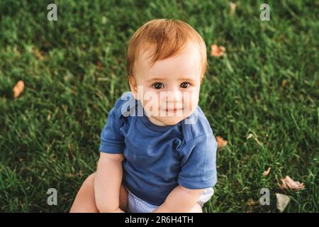 Portrait extérieur d'adorable bébé à tête rouge jouant dans le parc d'été, assis sur l'herbe verte, portant un body bleu Banque D'Images