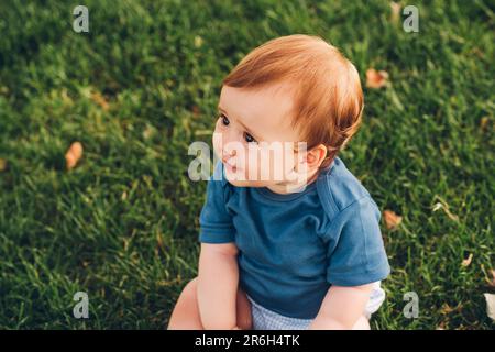 Portrait extérieur d'adorable bébé à tête rouge jouant dans le parc d'été, assis sur l'herbe verte, portant un body bleu Banque D'Images