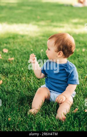 Portrait extérieur d'adorable bébé à tête rouge jouant dans le parc d'été, assis sur l'herbe verte, portant un body bleu Banque D'Images