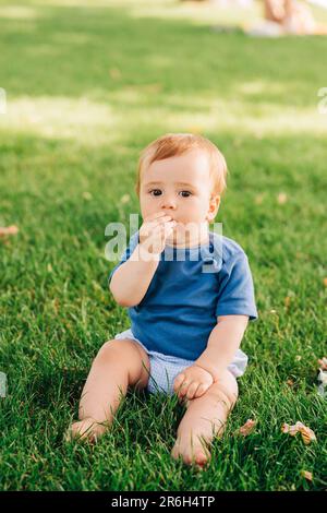 Portrait extérieur de bébé adorable à tête rouge jouant dans le parc d'été, assis sur l'herbe verte, portant un body bleu, tenant la main près de la bouche Banque D'Images