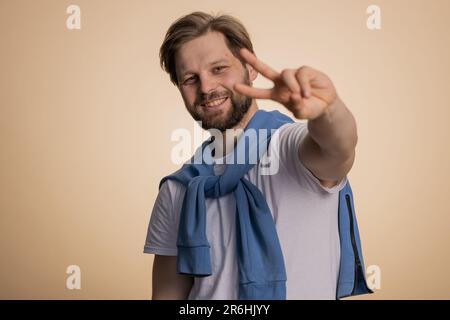 Jeune homme caucasien montrant le signe de la victoire, espérant le succès et la victoire, faisant le geste de la paix, souriant avec une expression aimable et optimiste. Beau gars barbu isolé seul sur fond beige studio Banque D'Images