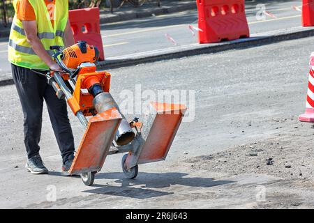 Un ouvrier de la route dans une cape réfléchissante orange déplace une foreuse à essence sur une partie réparée de la route par temps ensoleillé. Copier l'espace. Banque D'Images