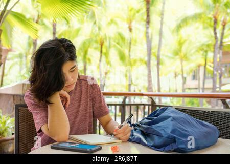 Jeune femme écrivant dans un carnet à l'extérieur dans un endroit tropical avec des palmiers en arrière-plan ensoleillé Banque D'Images