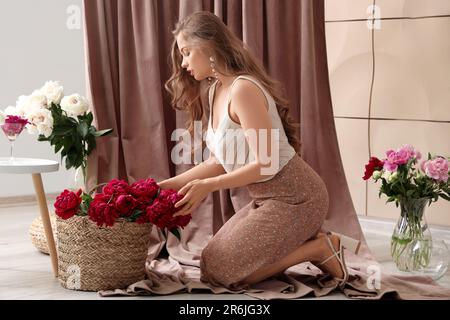 Belle jeune femme avec bouquets de fleurs de pivoine contre des rideaux dans la chambre Banque D'Images