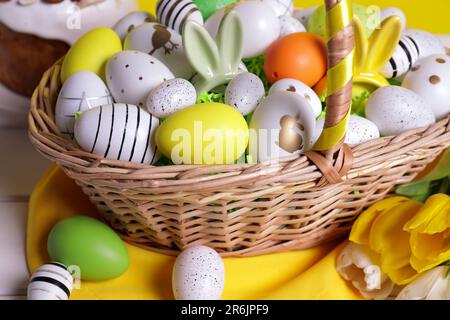 Panier en osier avec des œufs de Pâques décorés avec soin et de magnifiques tulipes sur table en bois blanc, en gros plan Banque D'Images