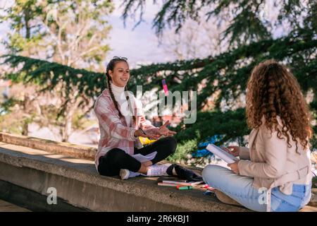 Deux filles d'école étudiant ensemble. Une jeune fille aux cheveux bouclés qui jette un marqueur à son ami. Ils sont assis sur une corniche murale Banque D'Images