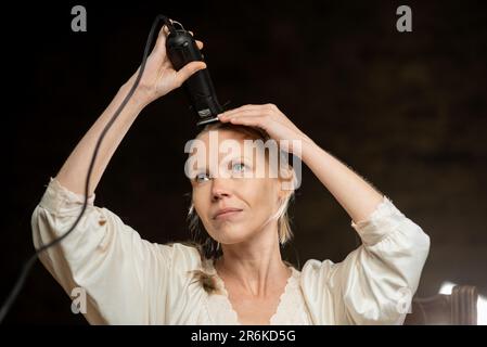 Une jeune femme d'âge moyen coupe tous ses cheveux sur la tête avec une tondeuse. arrière-plan sombre. Banque D'Images