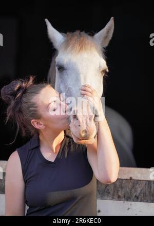 Jeune femme debout à côté du cheval arabe blanc reposant dans une boîte en bois écuries, l'embrassant les yeux fermés, le soleil brille sur eux, gros plan détail Banque D'Images