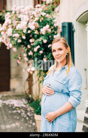 Portrait extérieur de femme enceinte élégante posant dans un jardin de roses, portant une robe en denim Banque D'Images