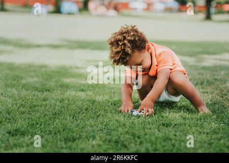 Adorable petit garçon africain jouant dehors avec des voitures jouets dans le parc sur l'herbe verte, portant un t-shirt de polo orange Banque D'Images