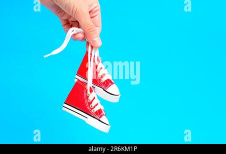 Gros plan d'une main tenant une paire de petites chaussures en toile rouge à haut sommet près des lacets sur un fond bleu vif. Mode pour enfants ou cadeau-rel Banque D'Images